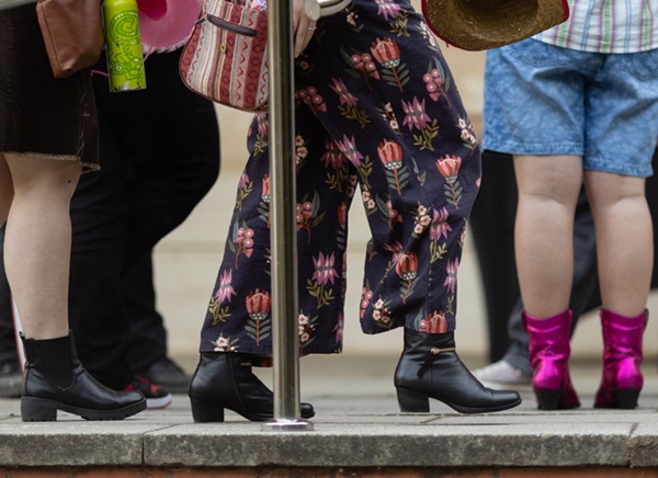 Image of a group of people from the waist down - most of them are wearing boots, one pair is bright pink.